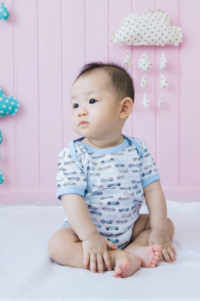 A baby boy sitting on a white blanket, wearing a blue and white patterned onesie, with a pink wall and a cloud-shaped mobile in the background.