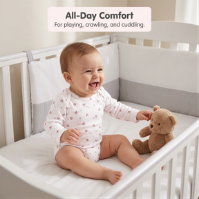 A smiling baby in a white onesie sits in a white crib, holding a brown teddy bear.