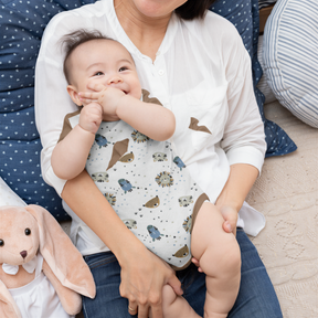 A woman is holding a baby who is chewing on her finger. The baby is wearing a white shirt with a pattern of animals. There is a stuffed animal, likely a bunny, next to the baby.