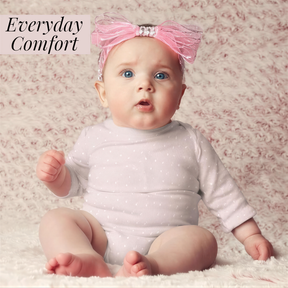A baby girl wearing a pink bow and a white polka dot outfit sits on a pink floral background, looking directly at the camera with a surprised expression.