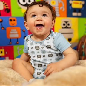 A smiling baby boy sitting on a bed, wearing a patterned onesie, with a colorful background featuring various cartoon robot characters.
