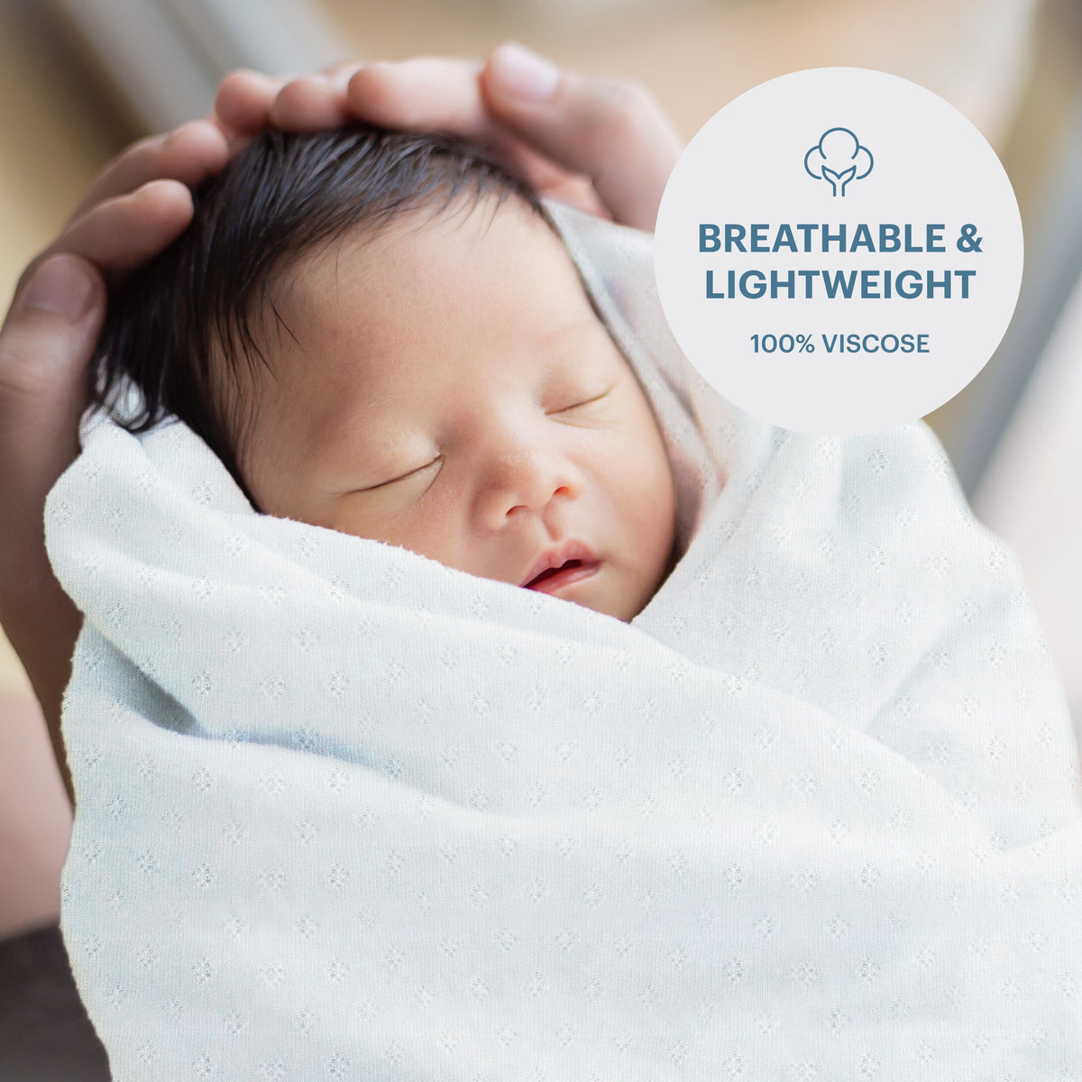 A newborn baby is wrapped in a soft white blanket, with hands gently cradling the baby's head.