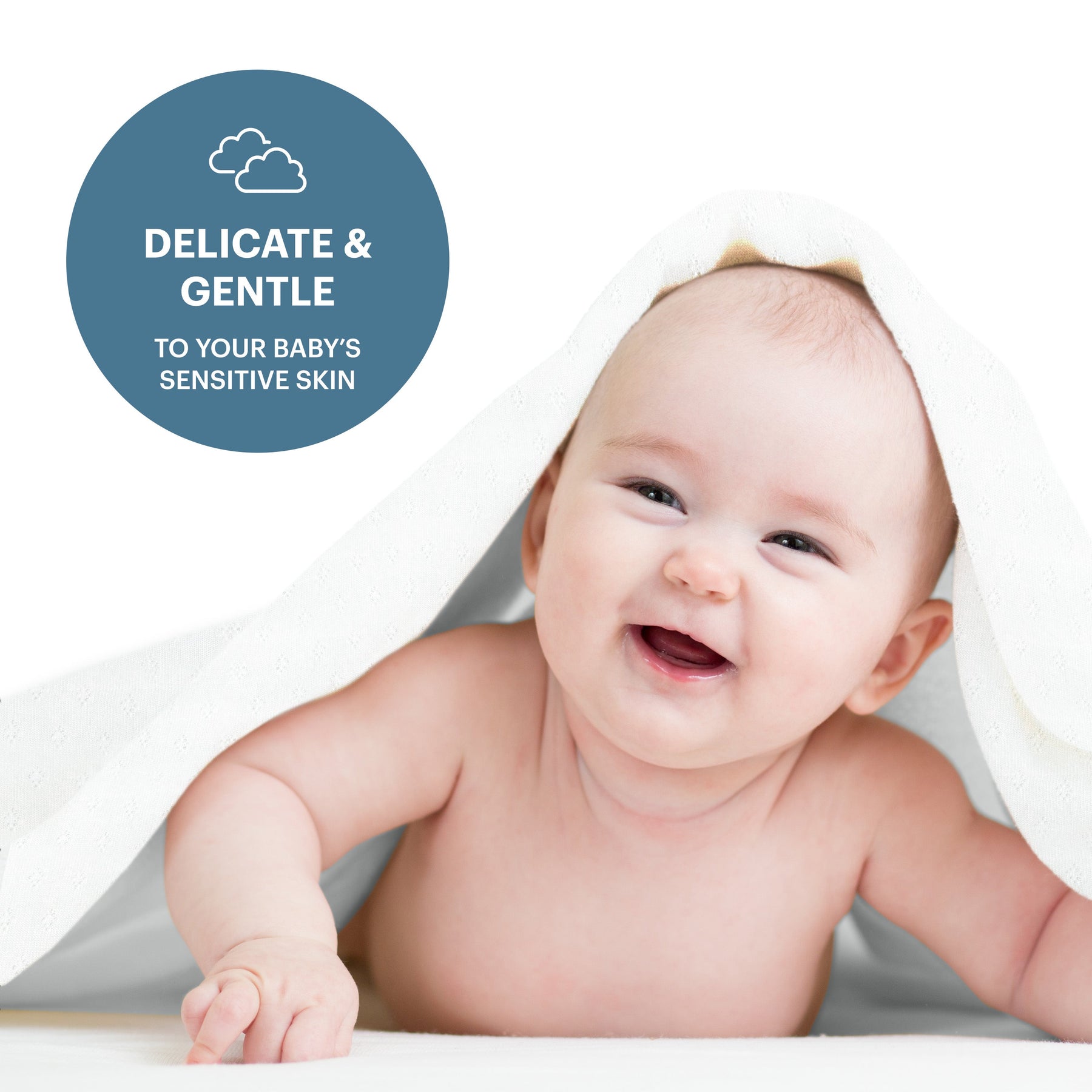 A smiling baby lying on a white surface, partially covered by a white towel.