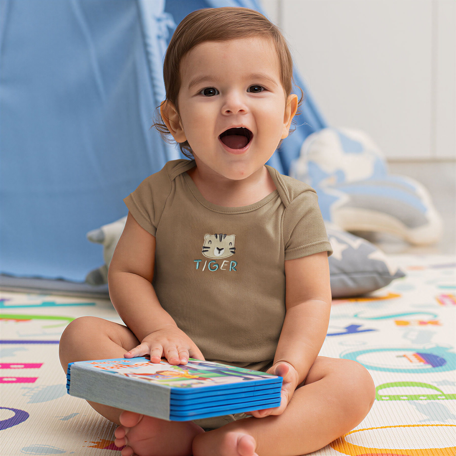 A smiling baby boy sitting on the floor, holding a book and looking at the camera.