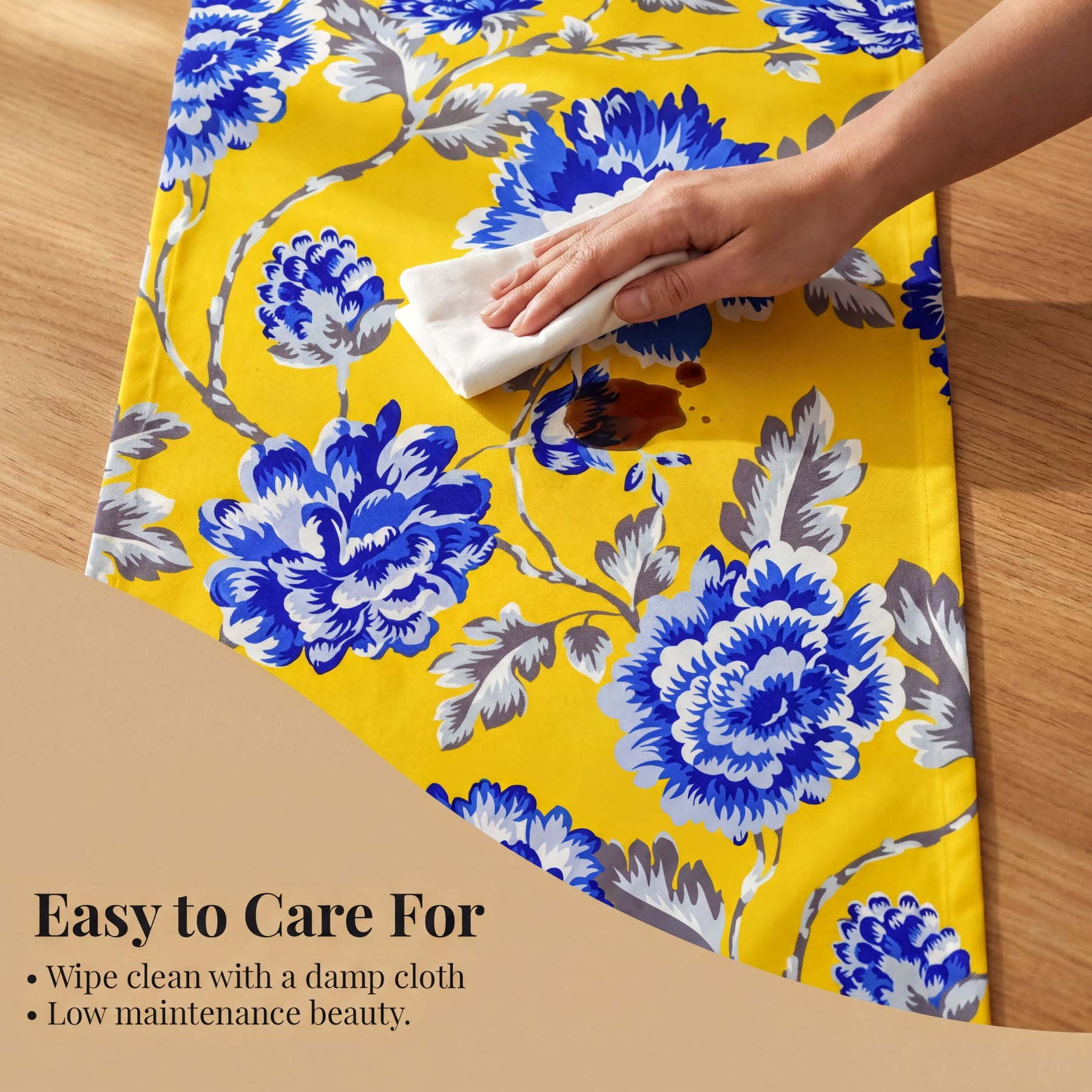 A hand is wiping a yellow and blue floral table runner with a damp cloth, as indicated by the text "Easy to Care For" and "Wipe clean with a damp cloth Low maintenance beauty."