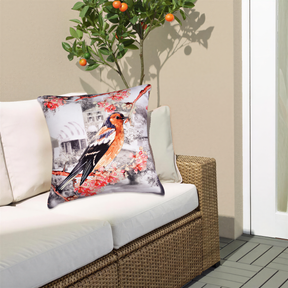 a white sofa with a decorative pillow featuring a colorful bird perched on a branch with flowers. The pillow is placed on the sofa, which is positioned against a wall with a wicker chair visible in the background.