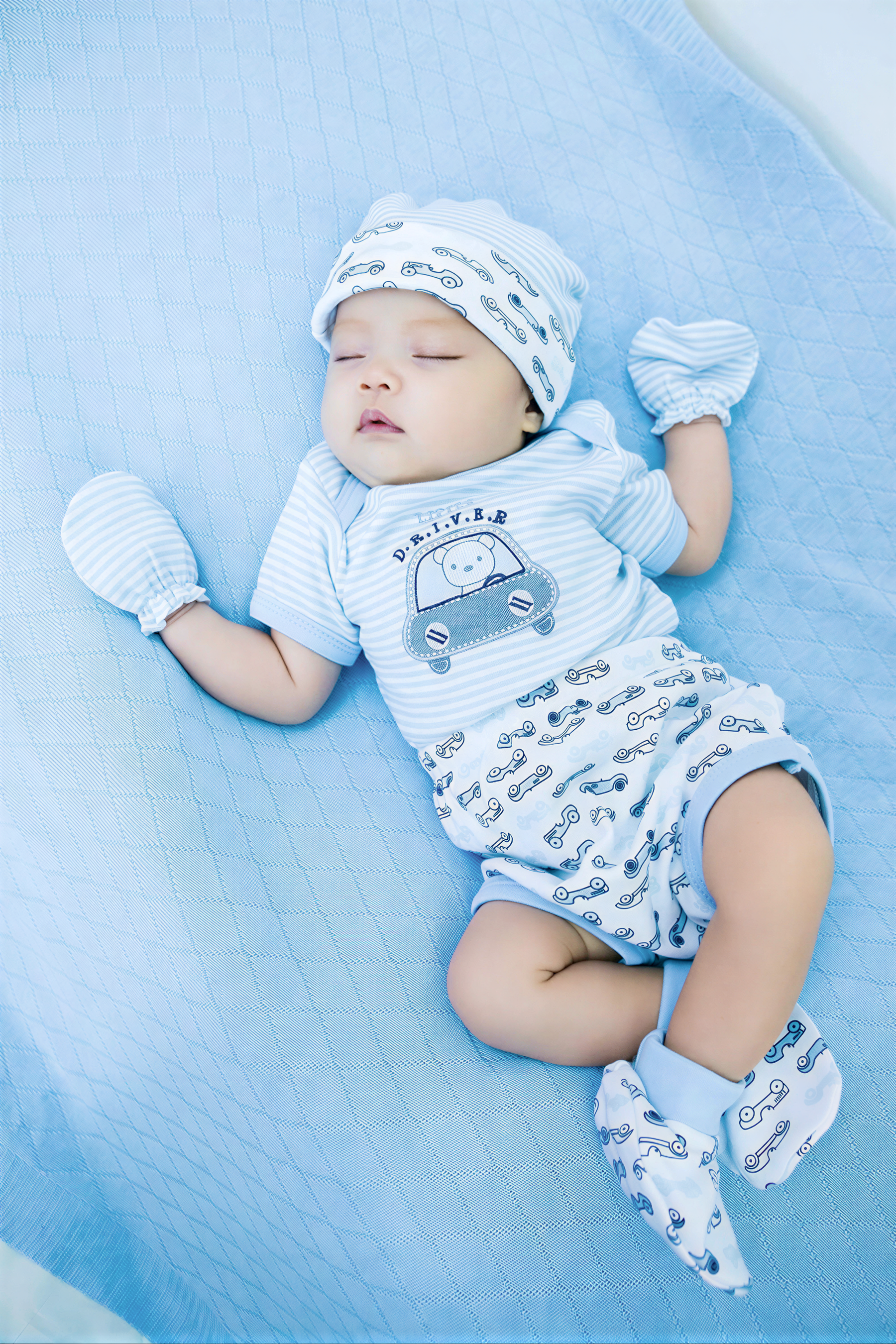 A sleeping baby wearing a blue outfit with a car design, lying on a blue blanket.