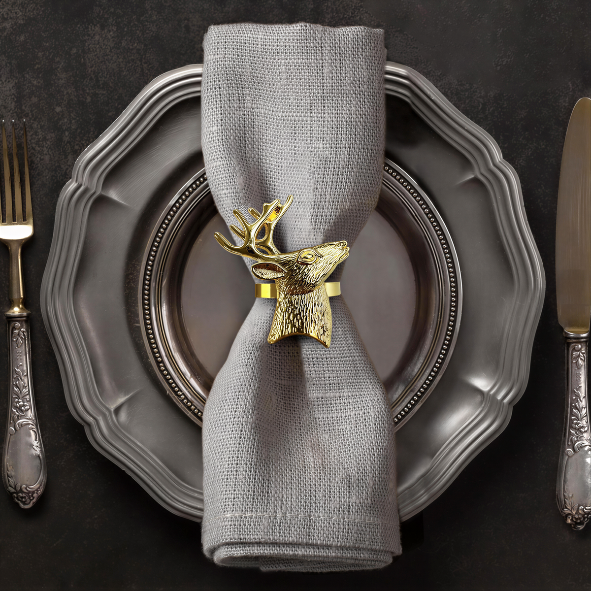A silver dinner plate with a decorative napkin ring featuring a gold deer head, accompanied by a fork and knife on a dark background.