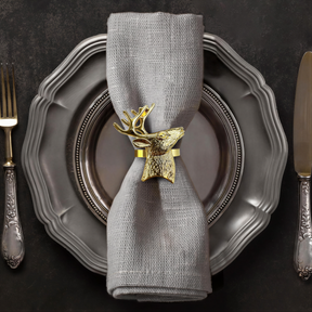 A silver dinner plate with a decorative napkin ring featuring a gold deer head, accompanied by a fork and knife on a dark background.