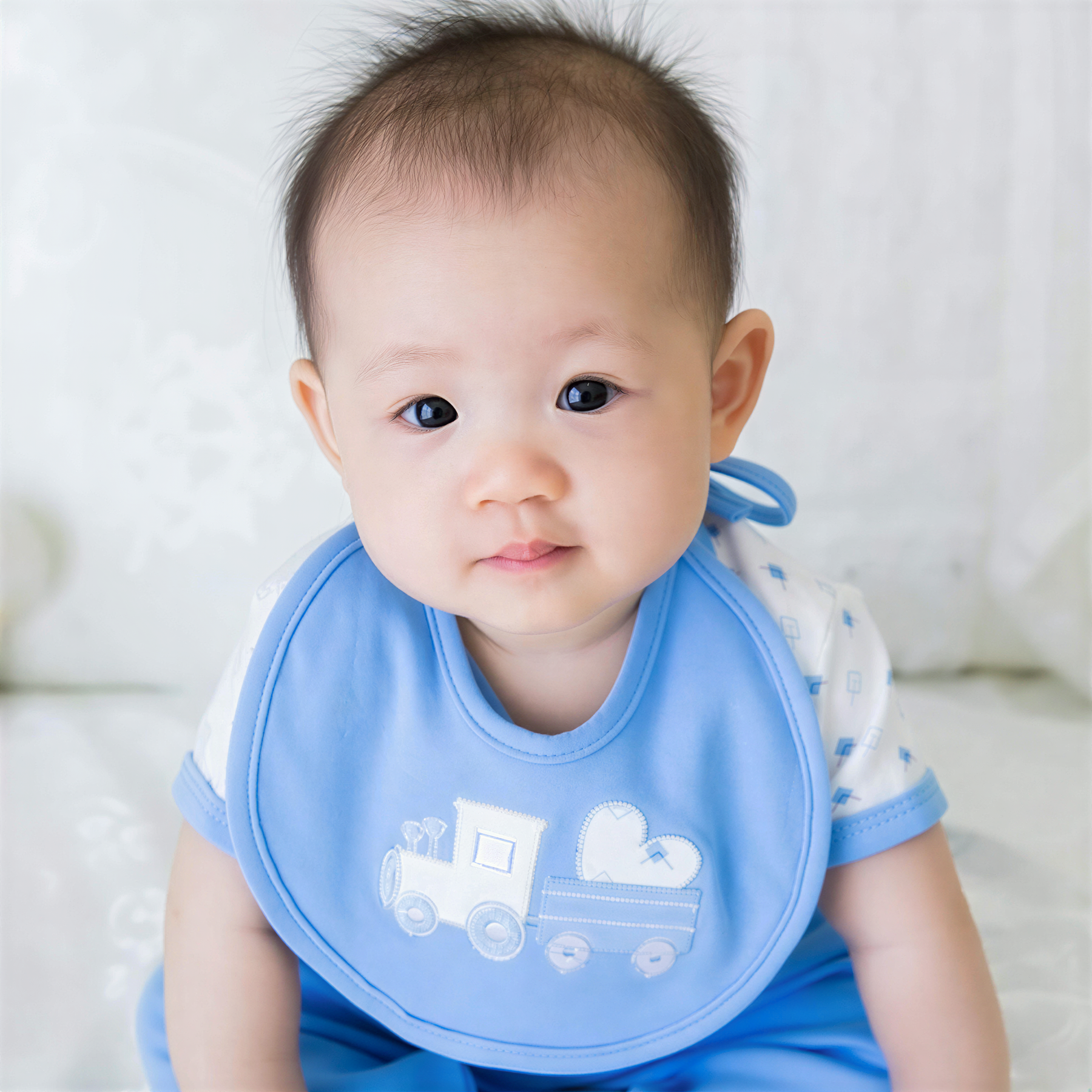 A baby boy wearing a blue bib with a train design, sitting on a white surface.