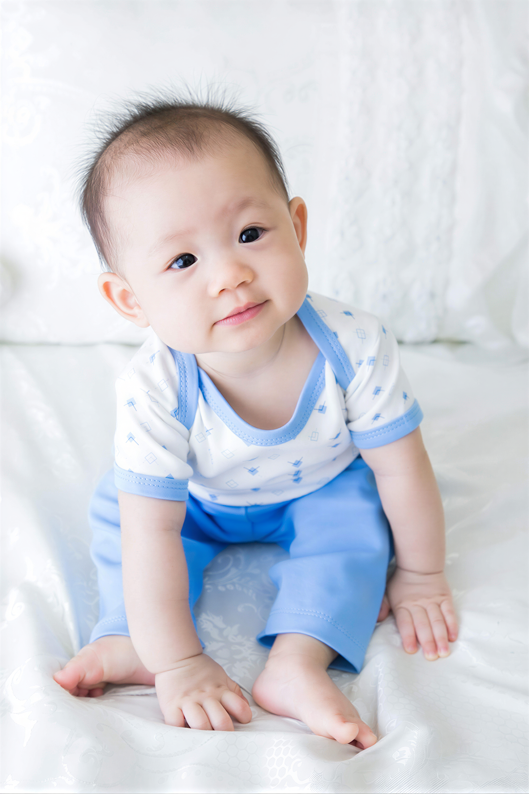 A smiling baby boy wearing a white and blue outfit sits on a white bedspread.
