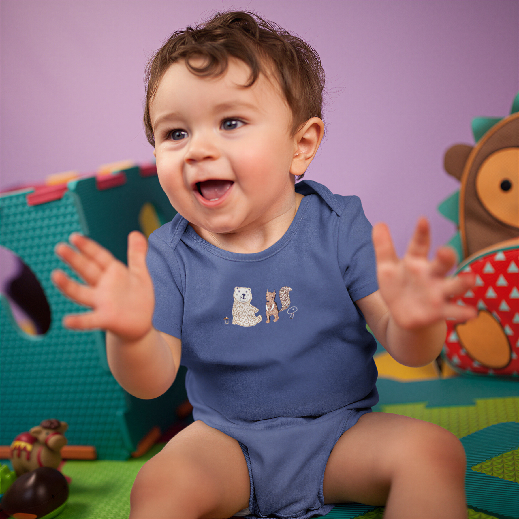 A happy baby boy wearing a blue onesie with a bear and a cat on it, sitting on a colorful play mat and smiling.