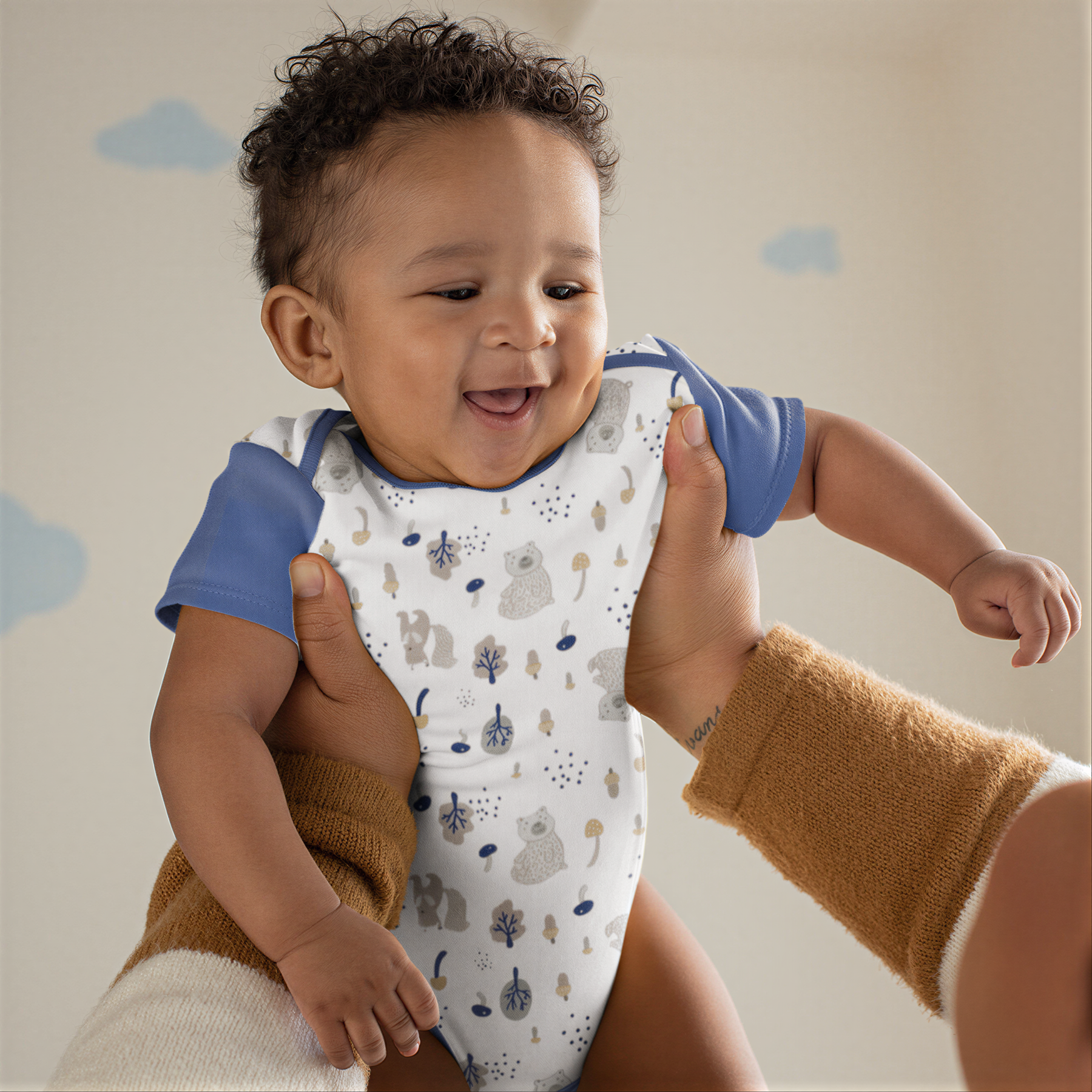 A smiling baby wearing a white onesie with a pattern of animals is being held by someone's arms.
