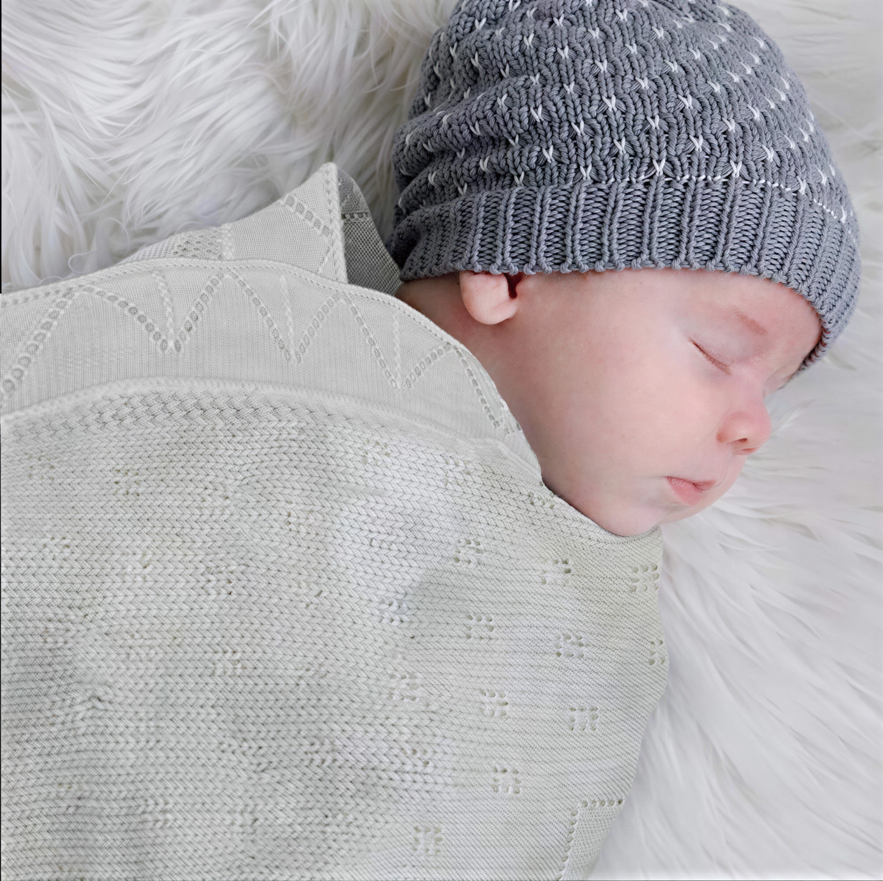 A sleeping baby wrapped in a white knitted blanket and wearing a gray knit hat.