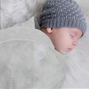 A sleeping baby wrapped in a white knitted blanket and wearing a gray knit hat.