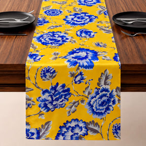 a table runner with a yellow floral print, placed on a wooden table. The runner is adorned with blue and white flowers, creating a vibrant and eye-catching design. The table is set with black plates and silverware, ready for a meal.