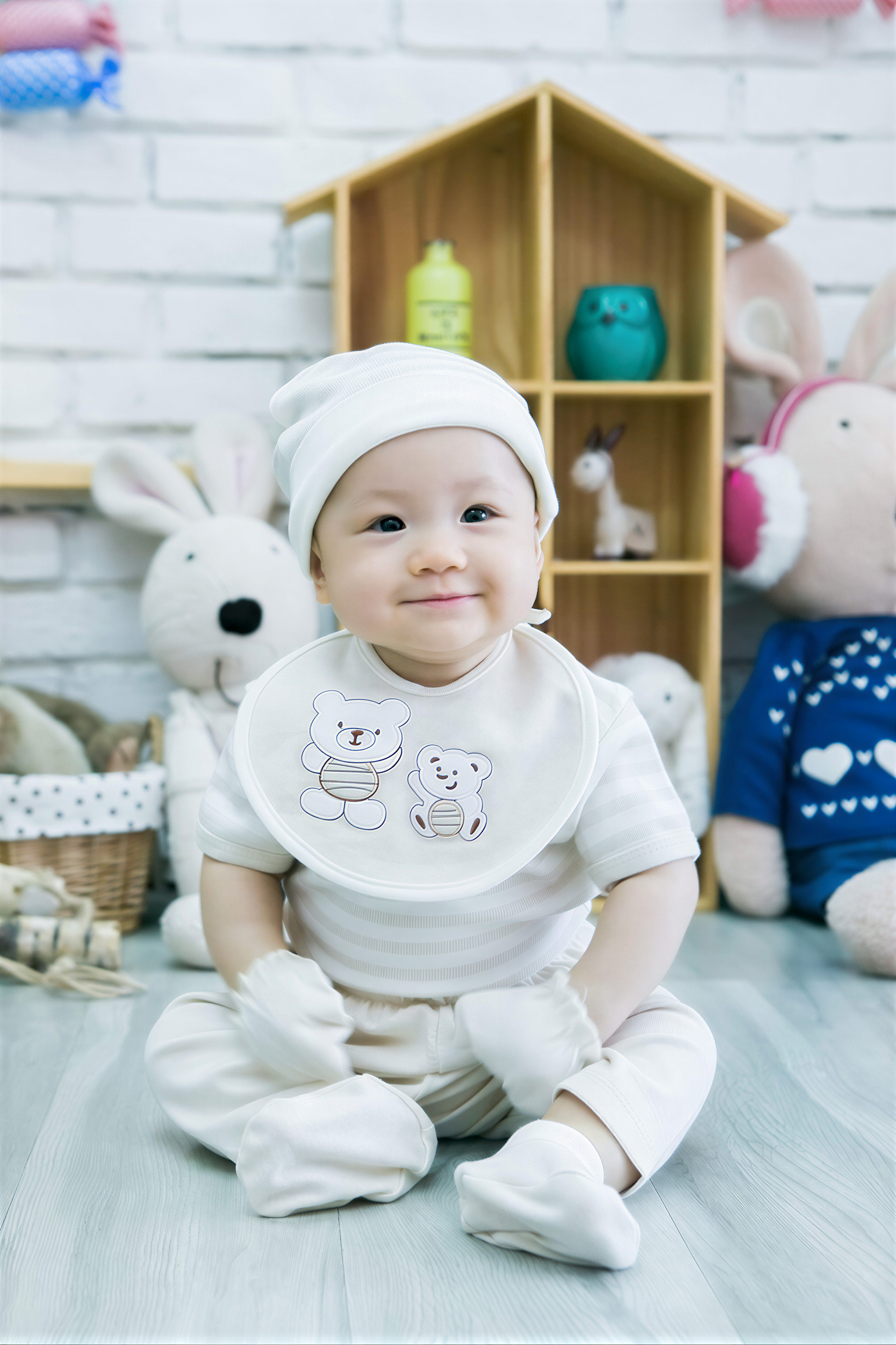 A smiling baby boy wearing a white outfit and bib sits on the floor in front of a wooden toy house, surrounded by stuffed animals.