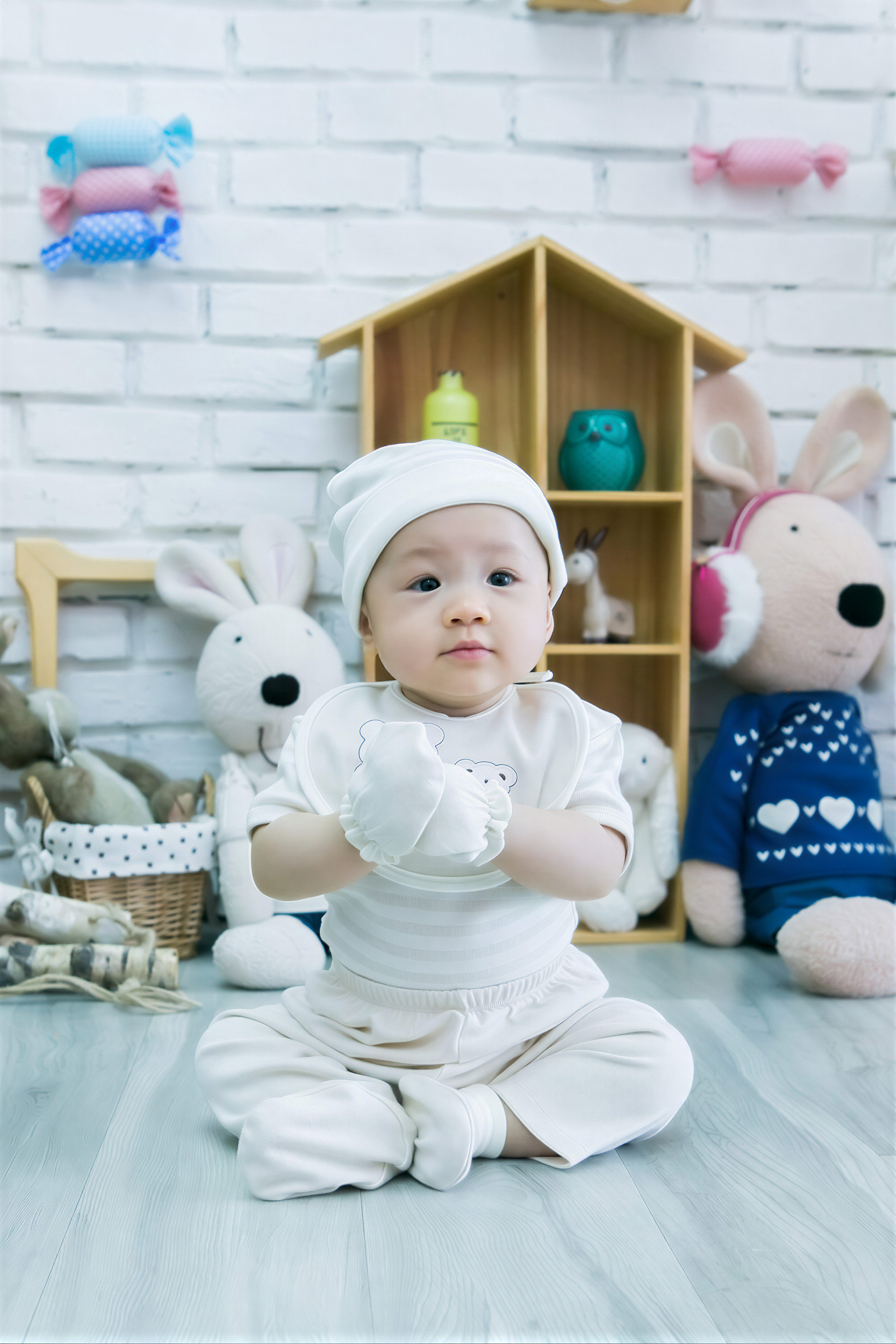 A baby sitting on the floor wearing a white outfit and hat, surrounded by stuffed animals and toys.