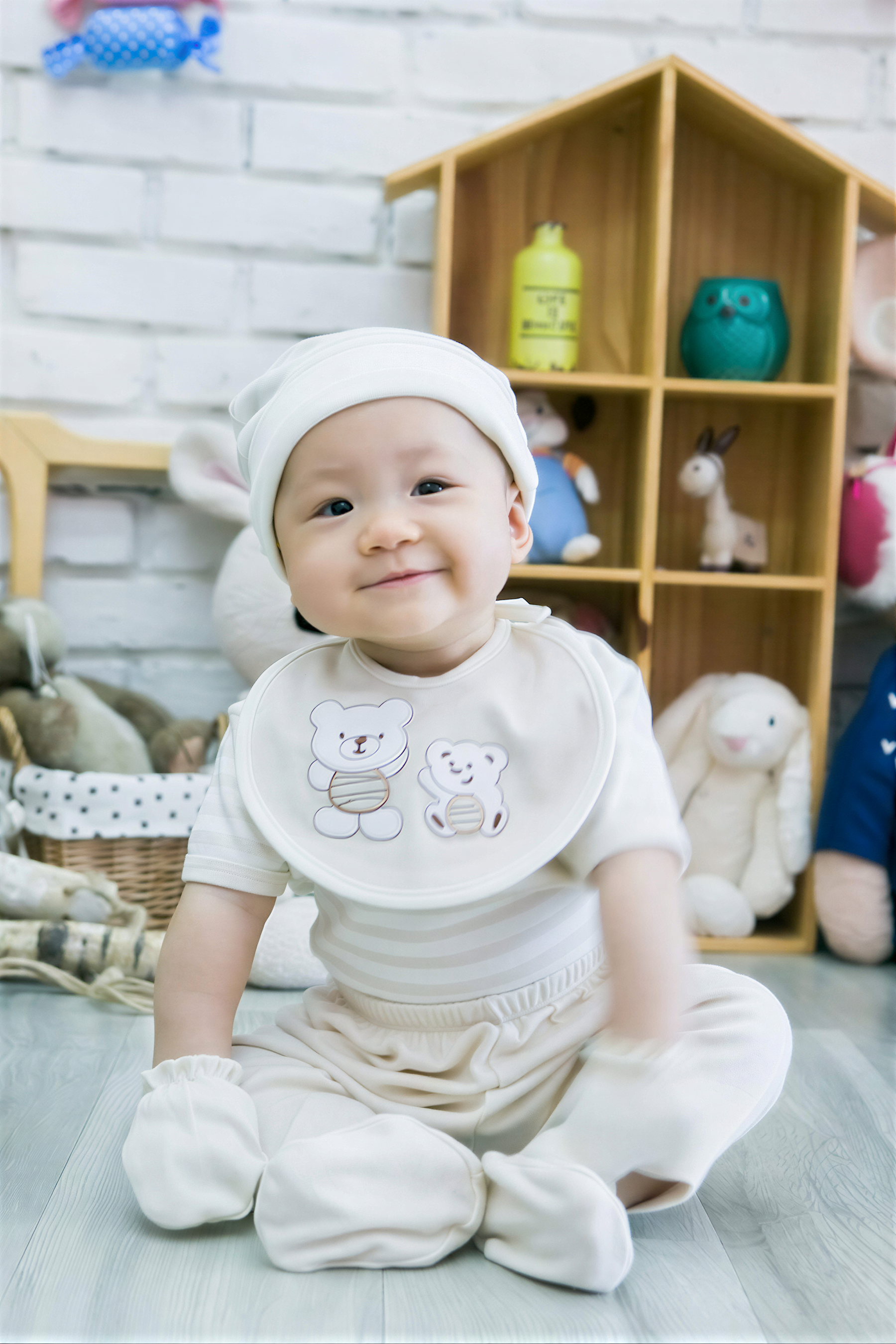 A smiling baby boy wearing a white outfit and bib sits on the floor in front of a wooden toy shelf filled with stuffed animals.