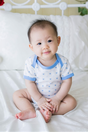 A smiling baby boy wearing a white onesie with blue polka dots sits on a white bed, looking directly at the camera.