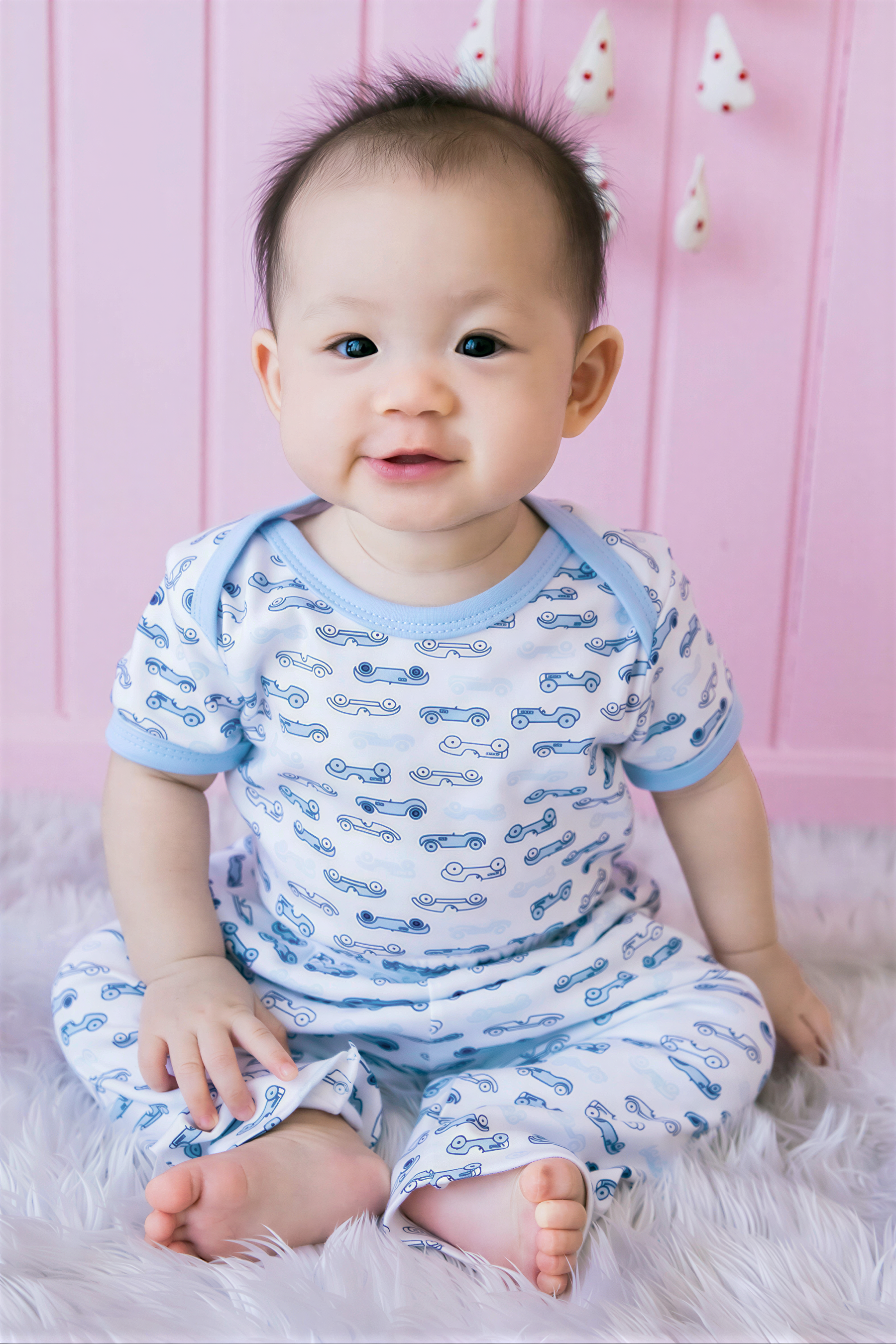 A smiling baby boy wearing a white onesie with blue car patterns sits on a pink background.