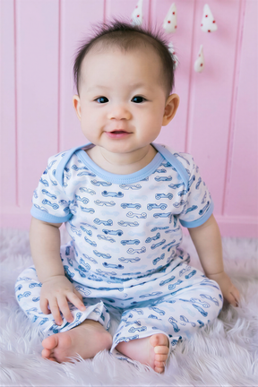 A smiling baby boy wearing a white onesie with blue car patterns sits on a pink background.