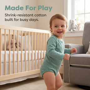 A smiling baby in a green polka dot onesie stands in a nursery, with a crib and a gray chair visible in the background.