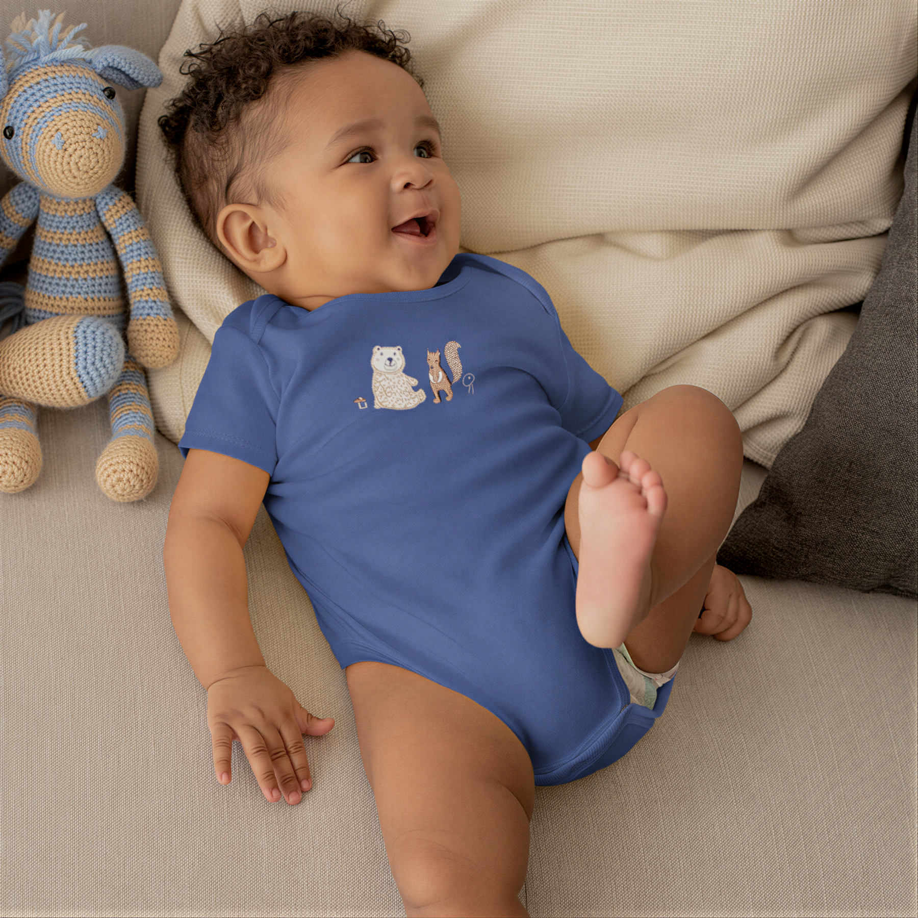 A smiling baby boy wearing a blue onesie with a bear and a squirrel on it, lying on a couch next to a stuffed animal.