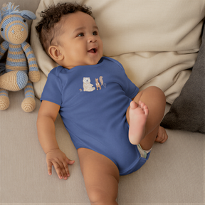 A smiling baby boy wearing a blue onesie with a bear and a squirrel on it, lying on a couch next to a stuffed animal.