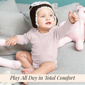A baby girl wearing a pink outfit and a knit hat is sitting on a gray couch, with a stuffed animal nearby.