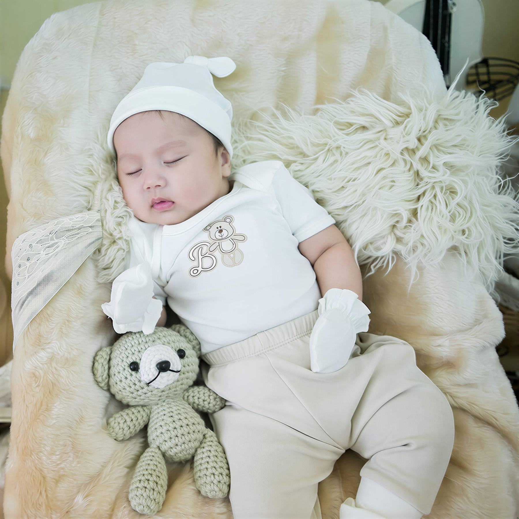 A baby is peacefully sleeping, wearing a white outfit with a teddy bear on their lap.