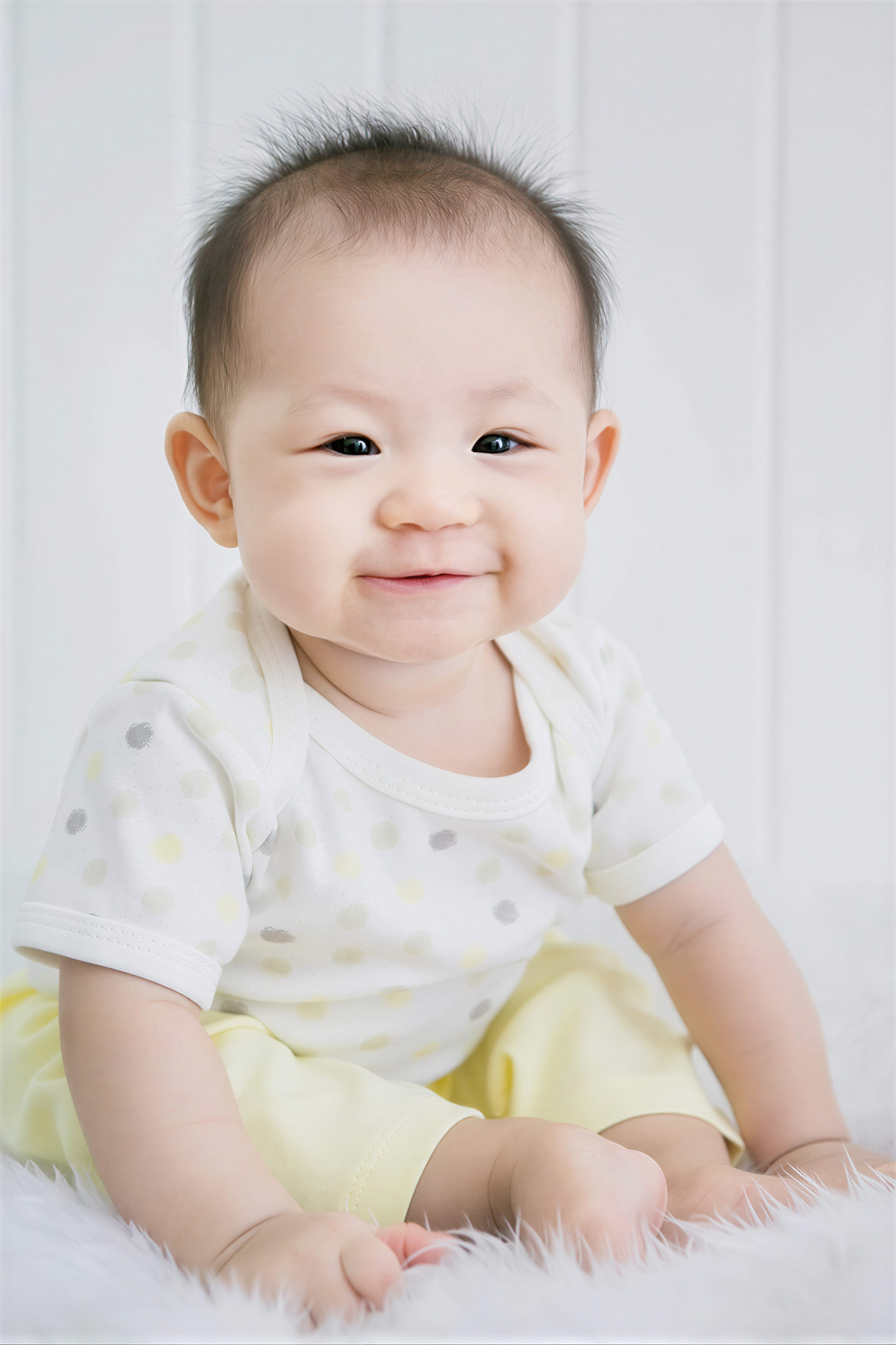 A smiling baby boy wearing a white polka dot shirt and yellow pants sits on a white fur rug.