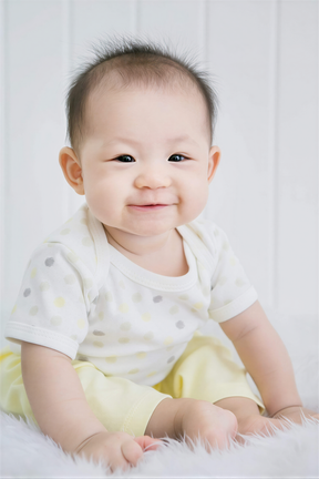 A smiling baby boy wearing a white polka dot shirt and yellow pants sits on a white fur rug.