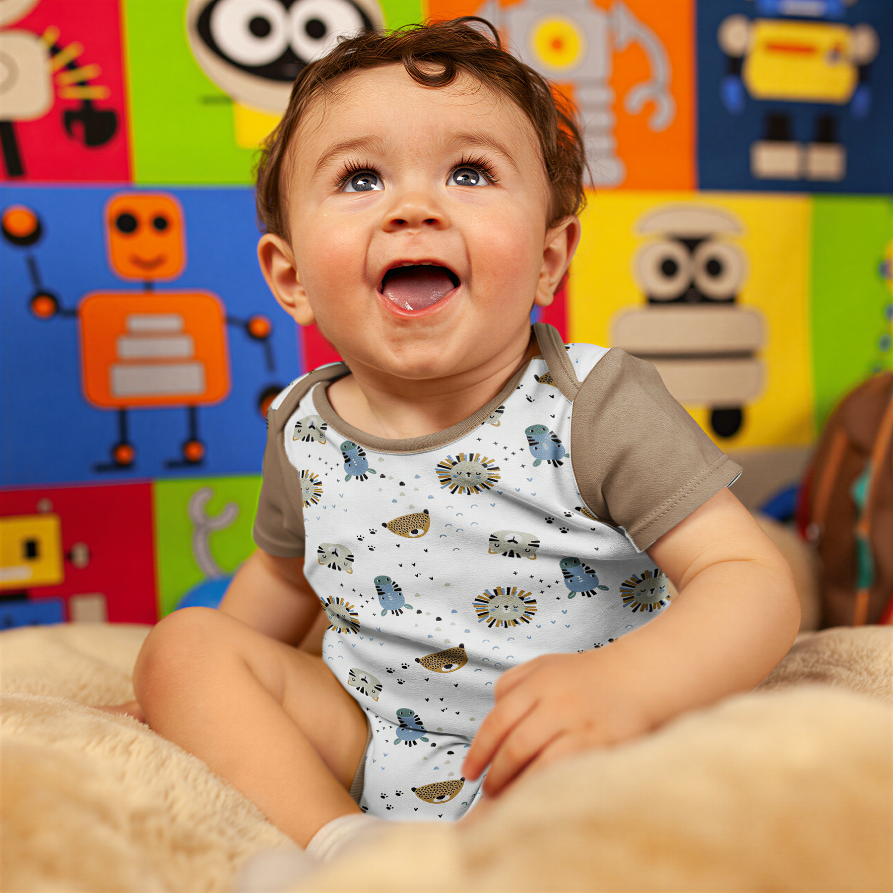 A smiling baby boy sitting on a bed, wearing a white onesie with animal designs.