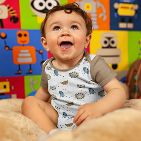A smiling baby boy sitting on a bed, wearing a white onesie with animal designs.