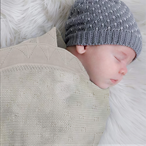 A sleeping baby wrapped in a beige knitted blanket and wearing a gray knit hat.
