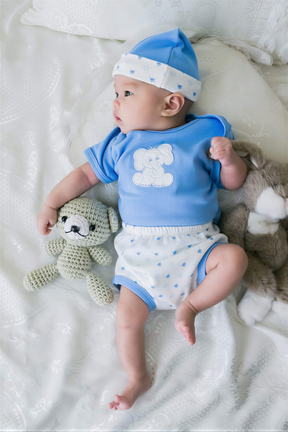 A baby wearing a blue outfit with a white polka dot pattern lies on a white bed, holding a small crocheted teddy bear.