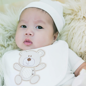 A baby wearing a white outfit with a teddy bear bib lying on a soft, fluffy surface.