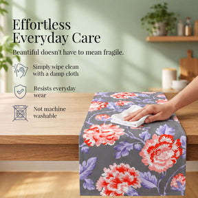 a person cleaning a table with a floral patterned table runner. The table runner has a floral design with red and purple flowers. The text above the image reads "Effortless Everyday Care" and below it, there are descriptions of the table runner's features, such as being easily wipeable, resistant to everyday wear, and not being machine washable.