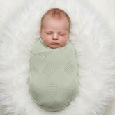 A newborn baby is peacefully sleeping, swaddled in a light green blanket, lying on a white fur rug.