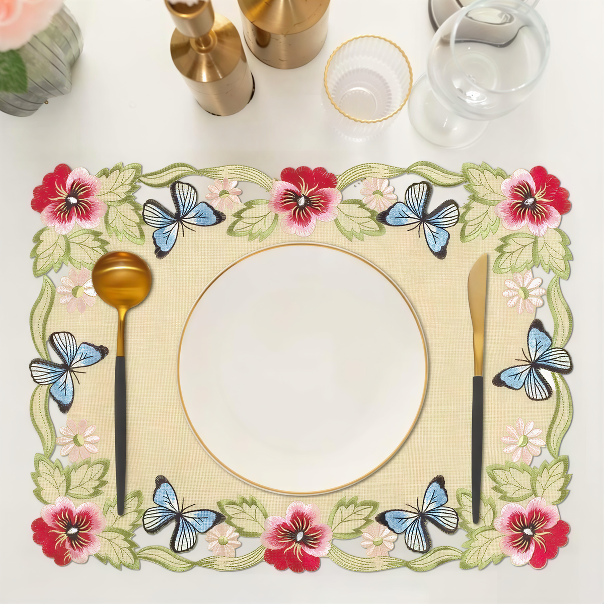 a table setting with a floral place mat, a white plate, a gold spoon, a gold knife, and a glass of water.