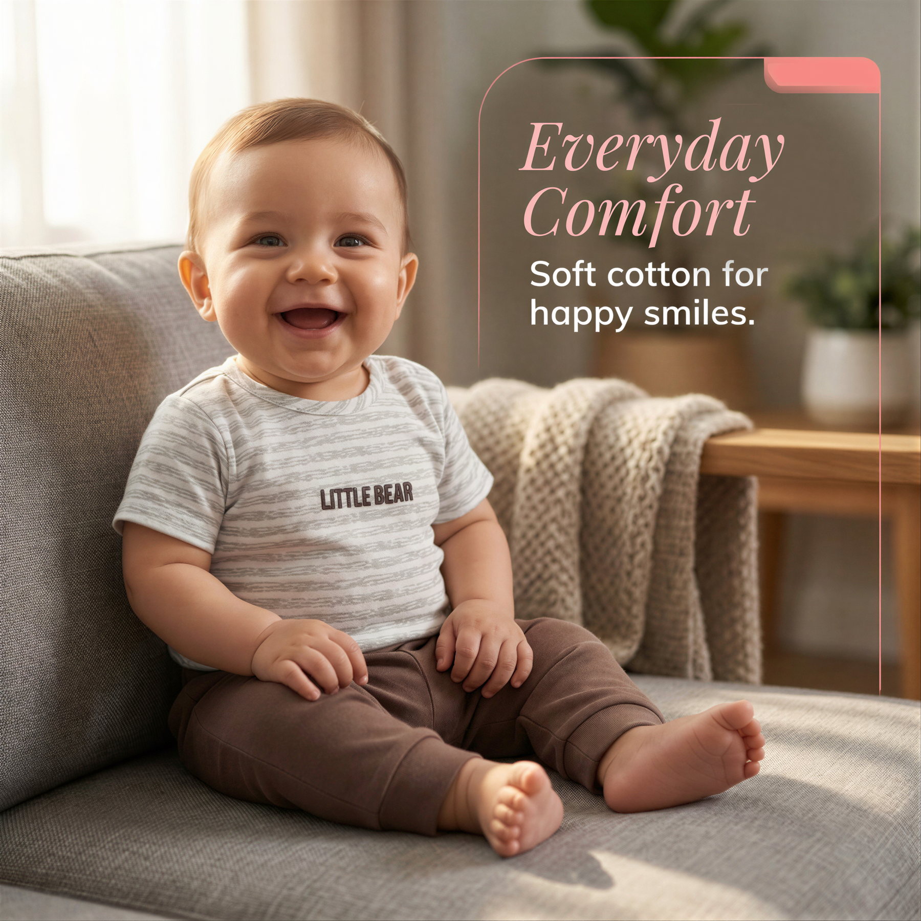 A smiling baby wearing a striped shirt and brown pants sits on a gray couch, with a potted plant visible in the background.