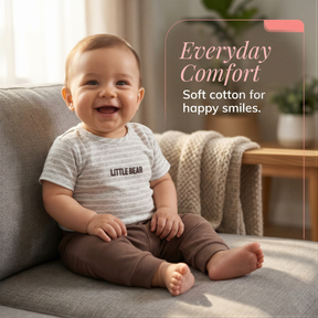 A smiling baby wearing a striped shirt and brown pants sits on a gray couch, with a potted plant visible in the background.