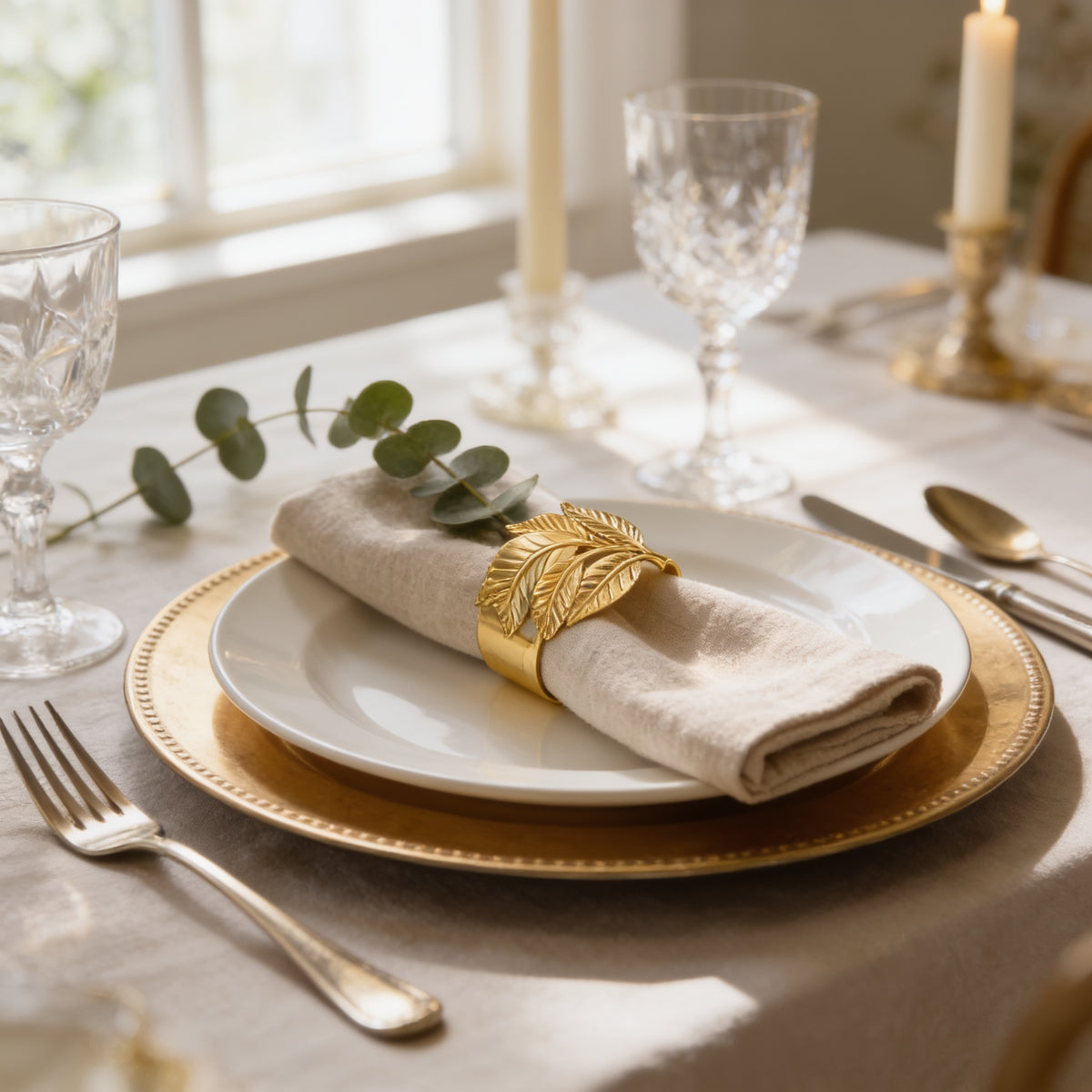 A table setting with a white plate, a folded napkin with a gold leaf ring, and a glass candle holder with a candle.