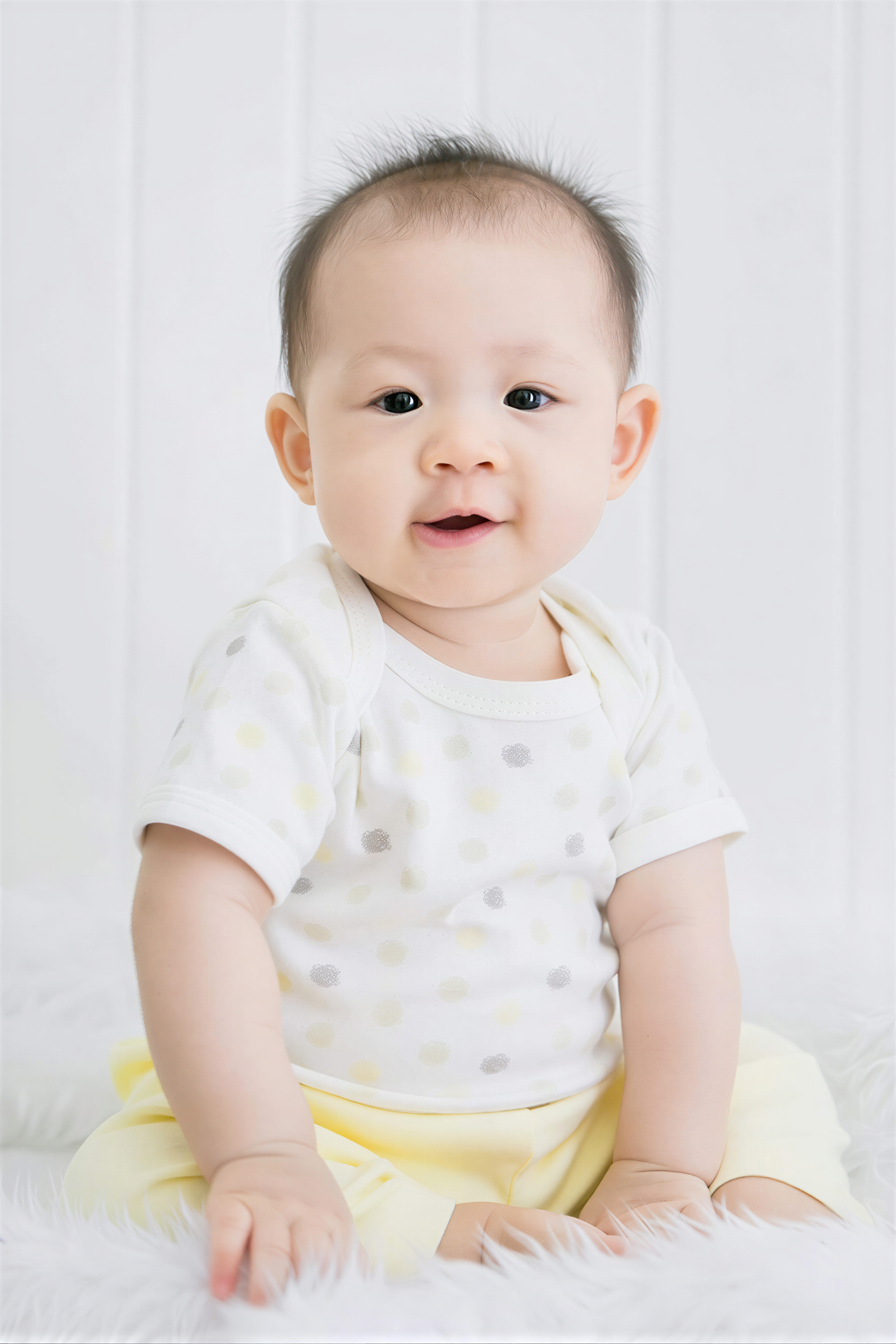 A smiling baby wearing a white polka dot shirt sits on a white fur rug against a white background.