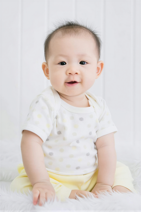 A smiling baby wearing a white polka dot shirt sits on a white fur rug against a white background.
