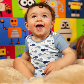 A smiling baby boy sitting on a bed, wearing a white and blue patterned onesie.