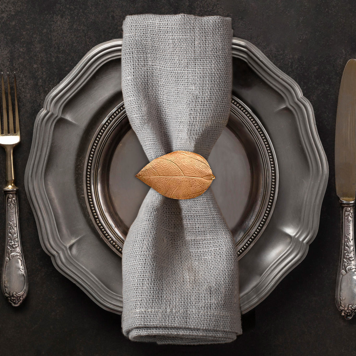 A silver dinner plate with a napkin ring featuring a leaf design, accompanied by a fork and knife on a dark background.