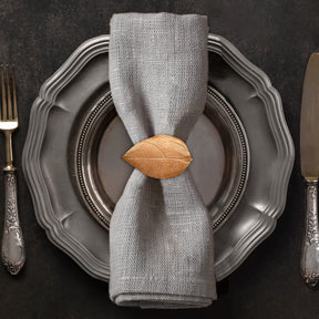 A silver dinner plate with a napkin ring featuring a leaf design, accompanied by a fork and knife on a dark background.