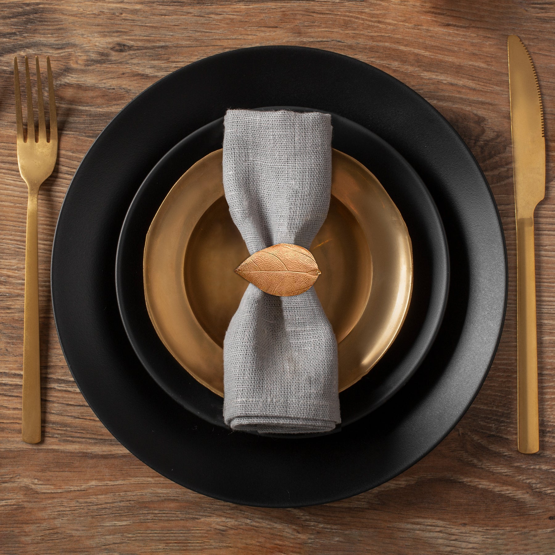 A black plate with a gold rim, a gray napkin ring, and a gold fork and knife are arranged on a wooden table.